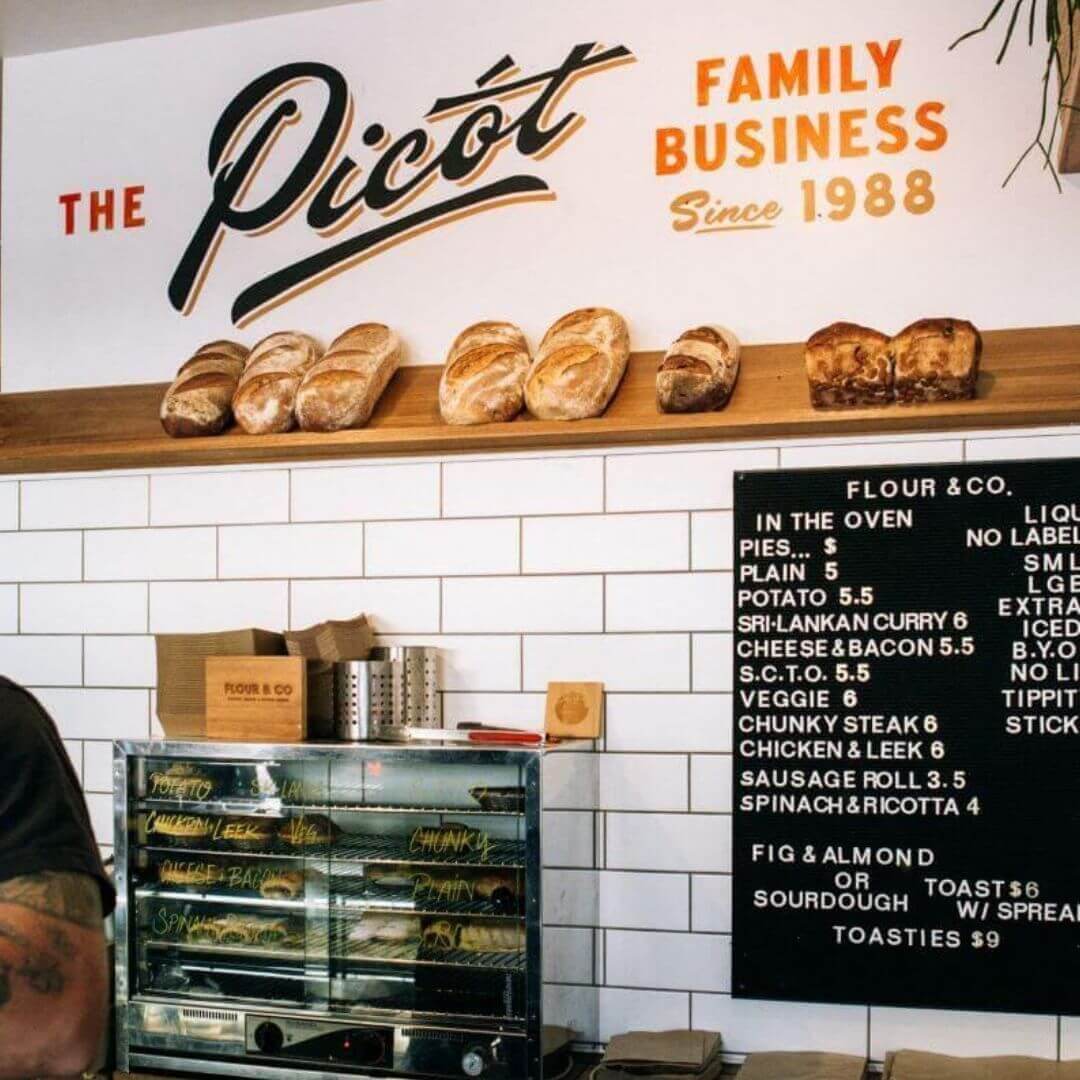 A cafe with a shelf of freshly baked bread, menu board, pie warmer and a sign that says "The Picot Family Business"